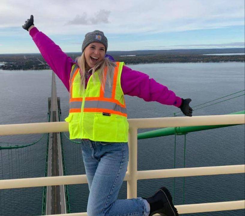 A woman in bright neon safety vest, hat, and gloves standing in front of a body of water